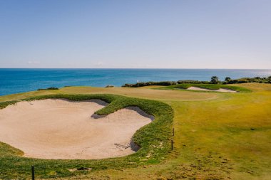 Endülüs, İspanya, 30 Ocak 2025 - Okyanus kıyısında açık mavi gökyüzü ve La Hacienda parkurunda sakin manzaralı bir golf sahasında kum sığınağı ve yeşil.