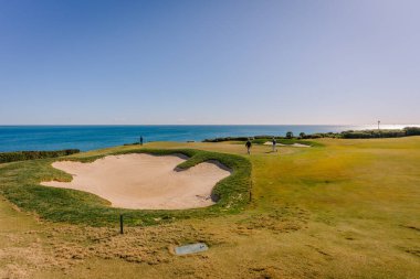Endülüs, İspanya, 30 Ocak 2025 - Golfçüler La Hacienda sahasında açık mavi gökyüzünün altında, okyanus kıyısındaki yeşil bir golf sahasında kumların yanında oynuyorlar.