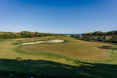 Endülüs, İspanya, 30 Ocak 2025 - Arka planda yeşil, kum sığınakları, ağaçlar ve okyanus olan golf sahası parlak mavi gökyüzünün altında, huzurlu bir manzara yaratıyor.