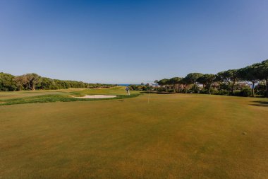 Andalucia, Spain, January 30, 2025 - Golf course with a putting green, sand bunker, trees, and a golfer in the distance under a sunny blue sky, creating a peaceful setting. clipart