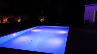 Gece aydınlık bir yüzme havuzu. Etrafı karanlık, ağaçlar ve arka planda dingin bir Buda heykeli ile çevrili..