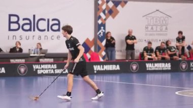 Valmiera, Letonya - 5 Şubat 2025 - biri kırmızı üniformalı, diğeri siyahlı iki florbol oyuncusu, bir maç sırasında koşuyorlar. Arka planda izleyiciler ve bir skorbord görünüyor.