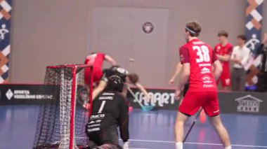 Valmiera, Letonya - 5 Şubat 2025 - Kırmızılı bir florbol oyuncusu kalenin yanında durup maçı izliyor. Bir kaleci kalenin önünde diz çöker ve diğer oyuncular sahada hareket eder..
