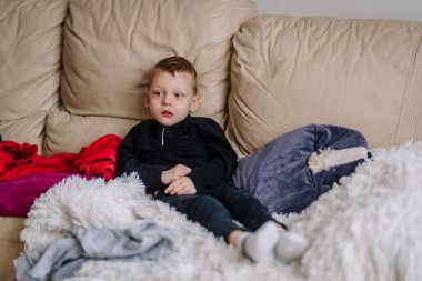 Bej bir kanepede oturan genç bir çocuk yumuşak yastıklar ve battaniyelerle çevrili, dikkatlice televizyon seyrediyor..