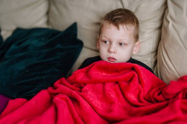 Hasta bir çocuk bej bir kanepede dinleniyor, parlak kırmızı bir battaniyeye sarılmış, yorgun ve sağlıksız görünüyor, kapalı bir odada yastıklarla çevrili..