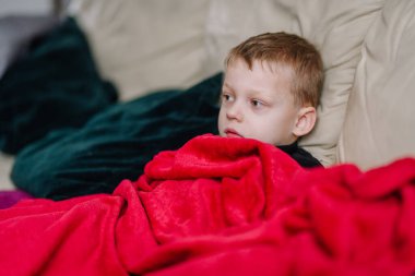 Hasta bir çocuk bej bir kanepede dinleniyor, parlak kırmızı bir battaniyeye sarılmış, yorgun ve sağlıksız görünüyor, kapalı bir odada yastıklarla çevrili..