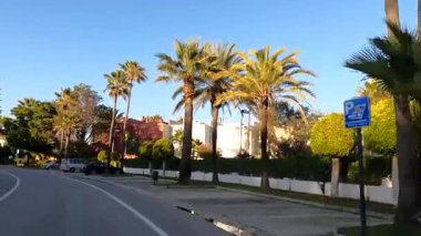 Endülüs, İspanya - 28 Şubat 2025 - Parlak mavi bir gökyüzü altında yemyeşil ve palmiye ağaçlarıyla çevrili İspanyol tarzı evlerin önünden geçiyor. Arabalar yol kenarına park edilmiş..