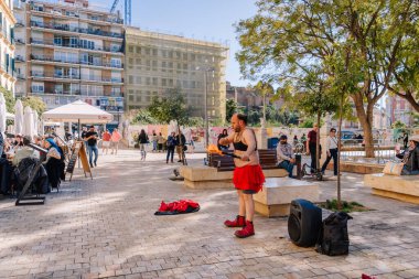 Endülüs, Malaga - 31 Ocak 2025 - Kırmızı etek ve siyah bluzlu bir sokak sanatçısı, kalabalık bir meydanda, kafeler ve ağaçlarla çevrili bir kalabalığı eğlendiriyor..