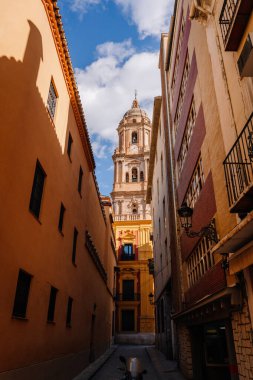 Andalucia, Malaga - January 31, 2025 - A narrow alleyway leading to the Malaga Cathedral, framed by traditional buildings under a bright blue sky with clouds. clipart