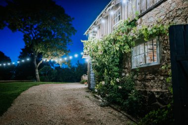 Cesis, Latvia - September 6, 2024 - Rustic stone building covered with vines, lit by string lights at night, with a gravel path leading to a serene garden under a deep blue sky. clipart