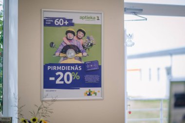 Ilukste, Letonya - 10 Eylül 2024 - Yaşlı bir çiftin scooter sürdüğü, pazartesi günleri yaşlılara% 20 indirim yapan bir eczane posteri (60 +).