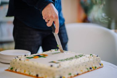 Ilukste, Letonya - 10 Eylül 2024 - Kutlama sırasında bıçakla beyaz kremalı pasta kesen bir kişi, yakın plakalı ve arka planı bulanık.