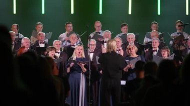 Valmiera, Letonya - 8 Şubat 2025 - Bir orkestra şefi tarafından yönetilen bir koro, seyirci tarafından izleniyor. Şarkıcılar nota kağıtlarını tutuyor ve sahnede renkli ışıklar var.. 