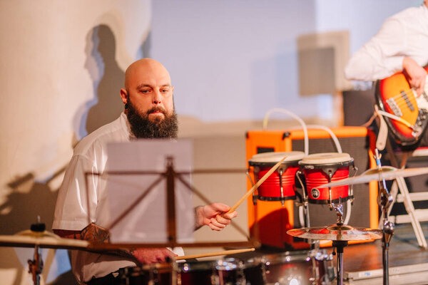 Валмиера, Латвия - 8 февраля 2025 - вечеринка 90-х годов. Бородатый барабанщик в белой рубашке выступает на сцене с ударной установкой, читая ноты, в то время как басист играет на заднем плане.