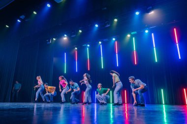 Valmiera, Letonya - 8 Şubat 2025 - 90 'ların partisi. Renkli gündelik kıyafetlerle dans eden bir grup genç erkek ve kadın dinamik ışıklandırmayla canlı bir sahnede dans ediyorlar..