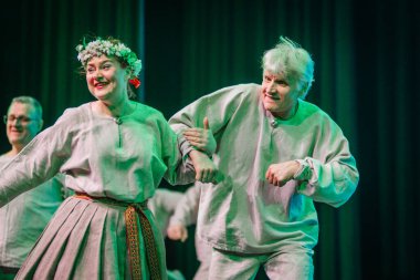 Valmiera, Letonya - 8 Şubat 2025 - 90 'ların partisi. Geleneksel beyaz ve gri folklor kostümlü bir grup kadın ve erkek mavi ve yeşil ışıklandırmalı bir sahnede neşeli bir dans ediyorlar..