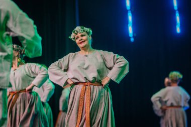 Valmiera, Letonya - 8 Şubat 2025 - 90 'ların partisi. Geleneksel beyaz ve gri folklor kostümlü bir grup kadın ve erkek mavi ve yeşil ışıklandırmalı bir sahnede neşeli bir dans ediyorlar..