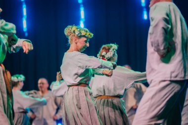 Valmiera, Letonya - 8 Şubat 2025 - 90 'ların partisi. Geleneksel beyaz ve gri folklor kostümlü bir grup kadın ve erkek mavi ve yeşil ışıklandırmalı bir sahnede neşeli bir dans ediyorlar..