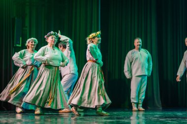 Valmiera, Letonya - 8 Şubat 2025 - 90 'ların partisi. Geleneksel beyaz ve gri folklor kostümlü bir grup kadın ve erkek mavi ve yeşil ışıklandırmalı bir sahnede neşeli bir dans ediyorlar..