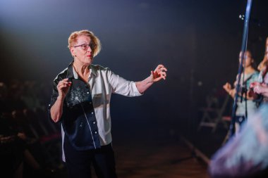Valmiera, Letonya - 8 Şubat 2025 - Bir orkestra şefi performans sırasında etkileyici el hareketleriyle bir koroya tutkuyla önderlik ediyor. Sahne, odaklanmış spot ışıklarıyla loş bir ışık altında. 90 'ların partisi