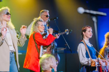 Valmiera, Letonya - 8 Şubat 2025 - Bir akordeonla şarkı söyleyen bir kadının, arka planda çalan müzisyenlerin ve geleneksel kıyafetli bir folk müzisyeninin yer aldığı canlı bir 90 'ların sahne performansı