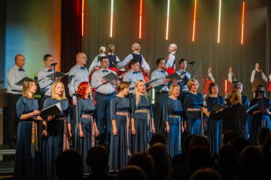 Valmiera, Letonya - 8 Şubat 2025 - Koro ve geleneksel dansçılar renkli ışıklar altında, bir orkestra şefinin önderliğinde, canlı bir kültürel 90 'ların parti atmosferi yaratarak sahnede gösteri yapıyorlar.