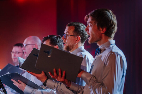 Валмиера, Латвия - 8 февраля 2025 - вечеринка 90-х годов. Группа певцов мужского хора выступает со страстью, держа в руках ноты, одетые в формальный наряд под теплым освещением сцены.