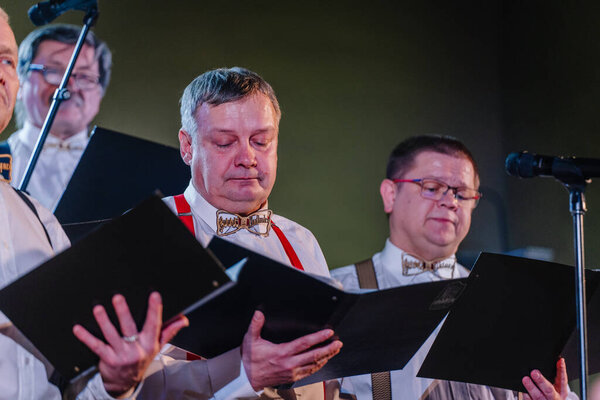 Валмиера, Латвия - 8 февраля 2025 - вечеринка 90-х годов. Пожилой человек поет в хоре, в очках и уникальной деревянной бабочке с музыкальными нотами, выступая на ярко освещенной сцене.