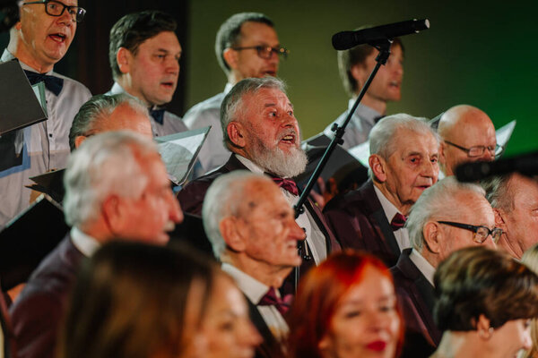 Валмиера, Латвия - 8 февраля 2025 - вечеринка 90-х годов. Хор выступает на сцене при драматическом освещении, одетый в формальный костюм, во главе с дирижером, при внимательном зрителе.