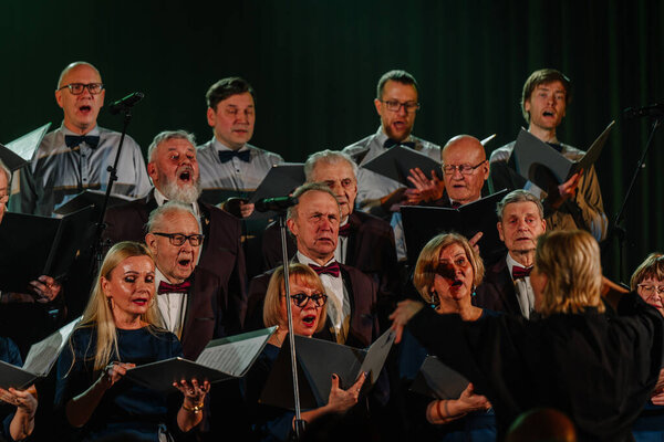 Валмиера, Латвия - 8 февраля 2025 - вечеринка 90-х годов. Хор выступает на сцене при драматическом освещении, одетый в формальный костюм, во главе с дирижером, при внимательном зрителе.
