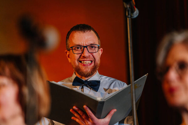 Валмиера, Латвия - 8 февраля 2025 года - Улыбающийся певец хора в очках и галстуке-бабочке держит на сцене песенник под теплым освещением, создавая радостную атмосферу вечеринки 90-х годов