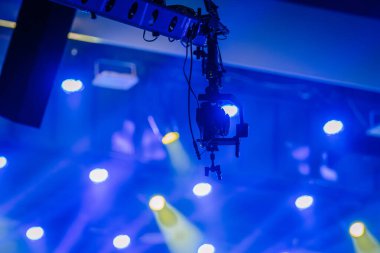 Valmiera, Letonya - 29 Aralık 2024 - Bir platformdan sarkan profesyonel bir kamera, arka planda mavi ve sarı sahne ışıklandırması olan canlı bir olayı kaydediyor..