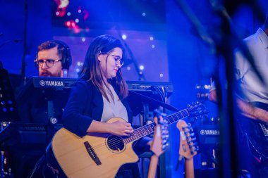Valmiera, Letonya - 29 Aralık 2024 - Yamaha etkinliği. Bir kadın gitarist, bir konser sırasında sahnede akustik gitar çalıyor, bir klavyeci ve diğer müzisyenler mavi sahne ışıkları altında şarkı söylüyorlar..
