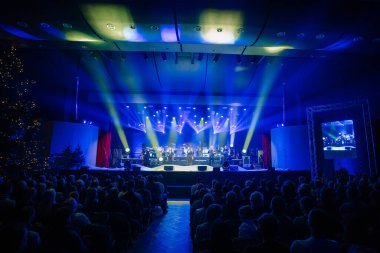 Valmiera, Letonya - 29 Aralık 2024 - Yamaha etkinliği. Parlak bir sahnede şarkı söyleyen şarkıcı ve müzisyenlerle dolu canlı bir konser. Seyirciler renkli ışıklandırmalar altında büyük bir salonda izliyor..