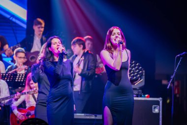 Valmiera, Letonya - 29 Aralık 2024 - Bir Yamaha etkinliğinde iki kadın şarkıcı canlı ışıklar altında arka planda gitar çalan bir grupla tutkuyla mikrofonlara şarkı söylüyor..