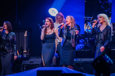 Valmiera, Letonya - 29 Aralık 2024 - Bir grup kadın şarkıcı Yamaha etkinliğinde sahne arkasında canlı sahne ışıkları altında canlı bir grupla tutkuyla mikrofonlara şarkı söylüyor..
