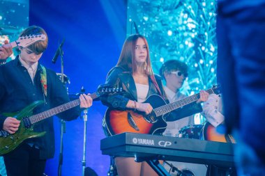 Valmiera, Letonya - 29 Aralık 2024 - Genç bir bayan gitarist Yamaha 'da canlı mavi sahne ışıklandırması altında diğer müzisyenlerle birlikte akustik gitar çalıyor..