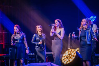 Valmiera, Letonya - 29 Aralık 2024 - Bir grup kadın vokal Yamaha etkinliğinde sahne alıyor, dramatik ışıklandırmayla aydınlatılıyor, ellerinde mikrofonlarla tutkuyla şarkı söylüyorlar..