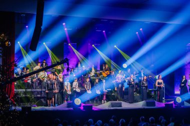 Valmiera, Letonya - 29 Aralık 2024 - Büyük Yamaha etkinliği canlı ışıklandırma efektleriyle aydınlatılmış bir sahnede şarkıcılar, müzisyenler ve perküsyonistlerle canlı performans sergiliyor.