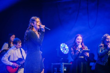 Valmiera, Letonya - 29 Aralık 2024 - Parlak elbiseli bir kadın şarkıcı, dramatik mavi ışık altında müzisyen ve vokalistlerin eşlik ettiği bir Yamaha etkinliğinde sahne aldı..