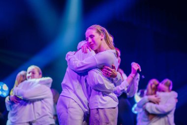 Valmiera, Letonya - 29 Aralık 2024 - Bir Yamaha etkinliğinde beyaz elbiseli iki sanatçı, dramatik mavi ışık altında derin duygu ve bağlantılarını ifade ederek sahnede kucaklaşıyorlar..