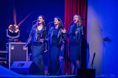 Valmiera, Letonya - 29 Aralık 2024 - Bir Yamaha etkinliğinde siyah elbiseli ve deri ceketli üç kadın şarkıcı dramatik ışıklandırma altında mikrofonlara tutkuyla şarkı söylüyorlar..