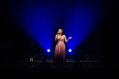Valmiera, Letonya - 29 Aralık 2024 - Bir Yamaha etkinliğinde akışkan pembe elbiseli bir kadın şarkıcı, dramatik mavi spot ışıklarıyla aydınlatılarak büyüleyici bir konser ortamı yarattı..