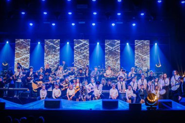 Valmiera, Letonya - 29 Aralık 2024 - Bir Yamaha etkinliğinde büyük bir gençlik orkestrası sahne alıyor, parlak mavi sahne ekranlarıyla gitar, klavye ve perküsyon çalıyor.