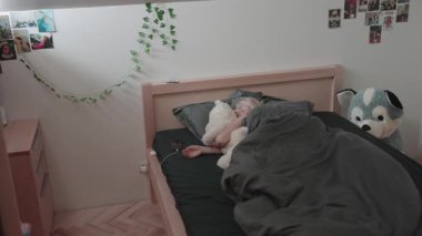 Bir kişi rahat bir yatak odasında uyuyor, siyah bir battaniyenin altında beyaz bir oyuncak ayıya sarılıyor. Pofuduk bir oyuncak yatağın yanında oturuyor ve yakınlarda bir telefon şarj oluyor..