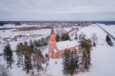 Velena, Letonya - 15 Şubat 2025 - Çan kulesine sahip kırmızı tuğlalı bir kilise, karlı kırsal bir arazide duruyor, her yer yemyeşil ağaçlarla, tarlalarla ve bulutlu bir gökyüzünün altındaki bir köyle çevrili..