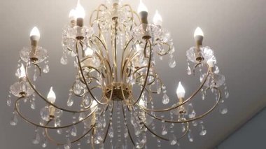 Altın metal kolları ve ışıldayan cam kolyeli lüks bir kristal avize, tavandan sarkan mum tarzı LED ampullerle aydınlatılmış..