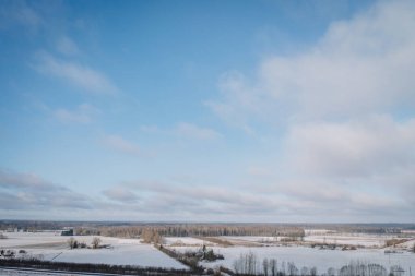Karla kaplı tarlaları ve açık mavi gökyüzünün altında dağılmış bulutlarla birlikte uzak ormanları olan sakin bir kış manzarası, kışın doğanın güzelliğini yansıtıyor..