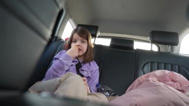 Mor ceketli genç bir kız arabanın arka koltuğunda oturur ve seyahat gereksinimleriyle çevrili bir şekilde emniyet kemeri takıp sosisli sandviç yer..