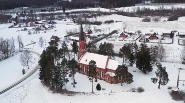 Velena, Letonya - 15 Şubat 2025 - Kırmızı tuğlalı bir kilise, ağaçlar, evler ve bulutlu bir gökyüzü altında uzanan uzun bir yol ile karla kaplı bir kırsal hava manzarası.